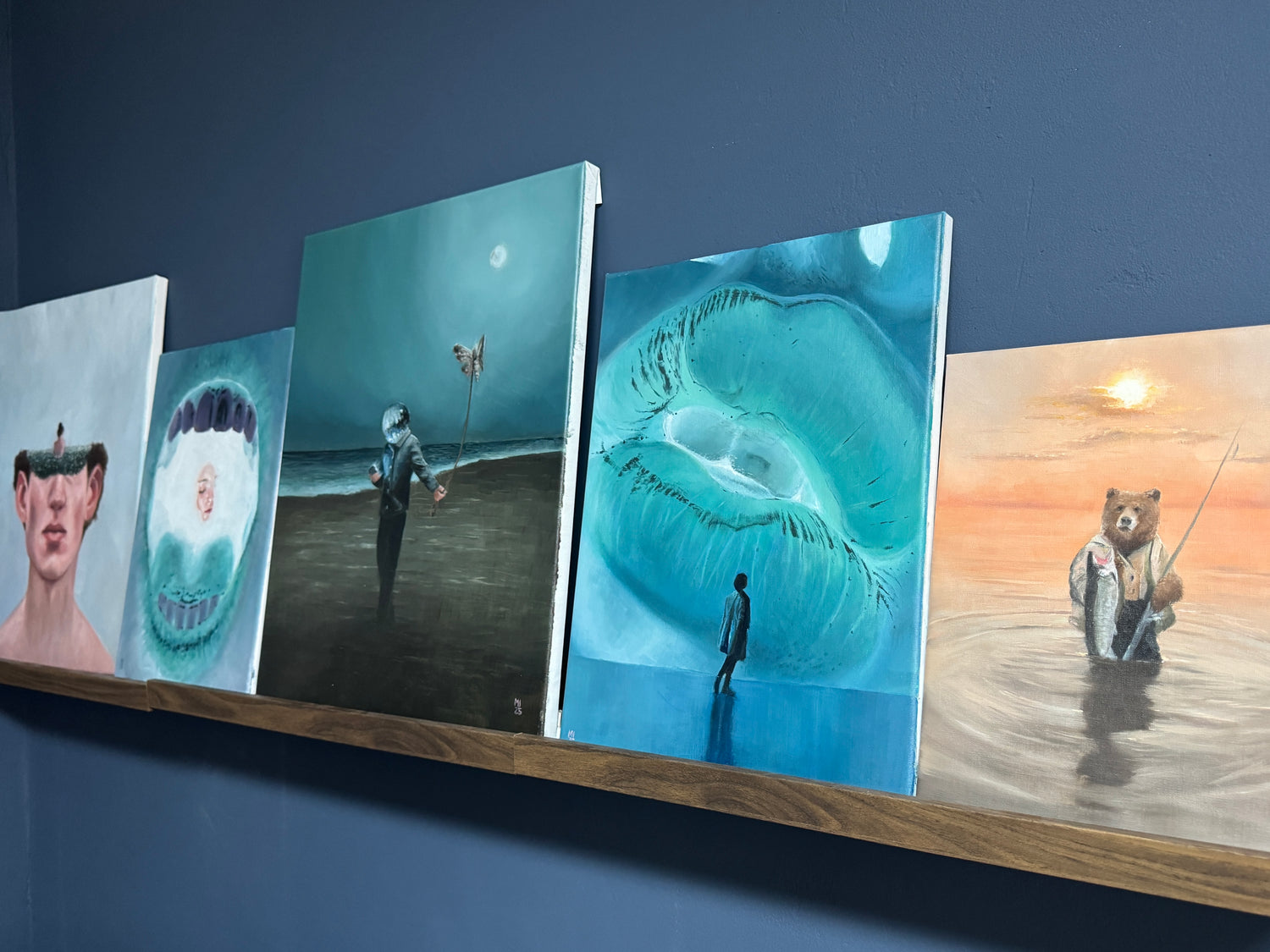 Five artistic paintings displayed on a wooden shelf against a blue wall.